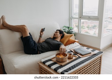 Asian man watch online tv and eating snack instead of working from home.