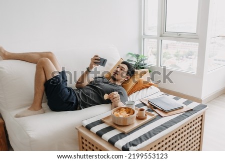 Asian man watch online tv and eating snack instead of working from home.