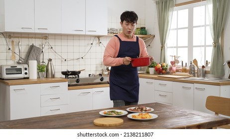 Asian Man Wearing Apron Setting The Table With Delicious Foods During The Day At Home. He Carries A Pot Of Hot Soup To The Table