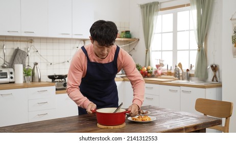 asian man wearing apron setting the table with delicious foods during the day at home. he carries a pot of hot soup to the table