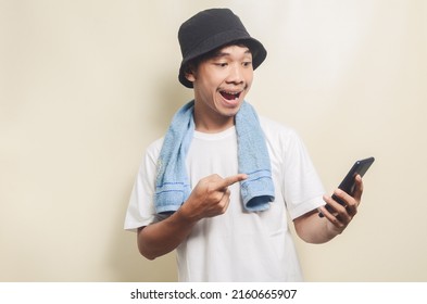 Asian Man Wearing Bright White T-shirt In Black Hat With Blue Towel Shocked To See His Phone On Isolated Background