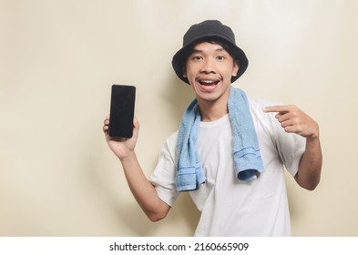 Asian Man Wearing Bright White T-shirt In Black Hat With Blue Towel Pointing At Phone Screen On Isolated Background