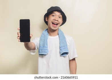 Asian Man Wearing Bright White T-shirt In Black Hat With Blue Towel Pointing At Phone Screen On Isolated Background