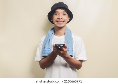 Asian Man Wearing Bright White T-shirt In Black Hat With Blue Towel Carrying Phone On Isolated Background