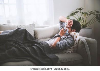 Asian Man Wearing A Hygienic Mask Is Not Feeling Well Resting On The Sofa In The House.