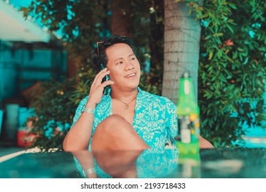 An Asian Man Wearing Light Blue Hawaiian Luau Polo Shirt Talking On His Phone In A Summer Garden Setting With Drink As Foreground.