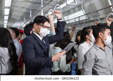 Asian Man Wearing Medical Face Mask Reading News Or Checking Work On Computer Tablet In Subway With Crowded People. New Normal Lifestyle During Coronavirus Pandemic