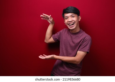 Asian Man Wearing Tshirt Over Red Background Gesturing With Hands Showing Big And Large Size Sign, Measure Symbol. Smiling Looking At The Camera. Measuring Concept.