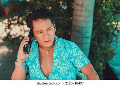 An Asian Man Wearing Unbuttoned Light Blue Hawaiian Luau Polo Shirt Usinghis Phone In A Summer Outdoor Garden