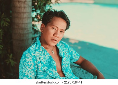 An Asian Man Wearing Unbuttoned Light Blue Hawaiian Luau Polo Shirt Posing And Looking At The Camera In A Summer Outdoor Garden