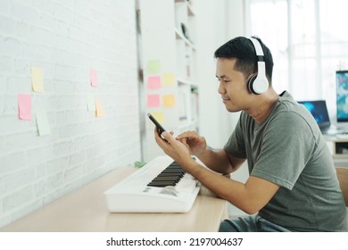 Asian Man Wearing White Headphones Play Piano And Sing A Song And Learning Online With Mobile Phone And Compose Writing Song Or Record Sound With A Microphone And Use Computer Recording Music Program