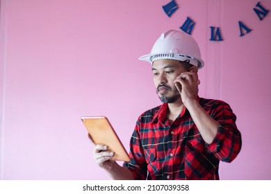 Asian Man Wearing White Helmet Wearing A Plaid Shirt Use Your Mobile Phone While Viewing The Screen On Your IPad.