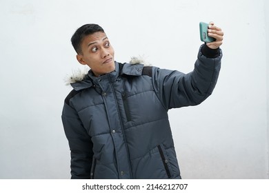 Asian man wearing winter jacket and holding smartphone taking selfie isolated on white background