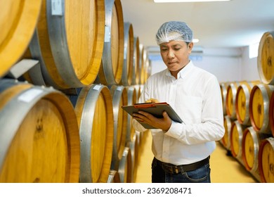 Asian man winemaker working and wine quality control in wine cellar with wooden barrels at wine factory. Winery manufacturing industry, brewery, alcohol liquor shop and winemaking concept. - Powered by Shutterstock