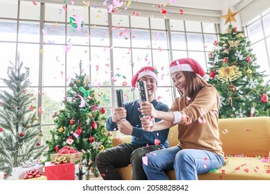 Asian Man And Woman With The Celebration Of Christmas In The Living Room. Asia Happy Couple Pull The Paper Fireworks And Gift Box With Christmas Tree In Santa Hats Sitting Couch At Home