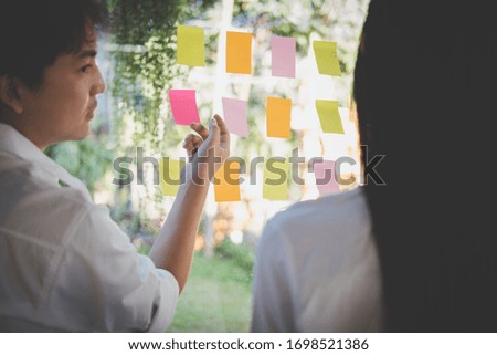 asian man & woman discussing creative idea with adhesive notes on glass wall at workplace. Sticky note paper reminder schedule at office. business, brainstorming, creativity concept