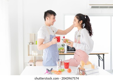 asian man and asian woman eating meal in the morning, they have breakfast in kitchen room, he holding hot coffee cup, she holding sandwich with hand, they feeling happy and smile, happiness honeymoon  - Powered by Shutterstock