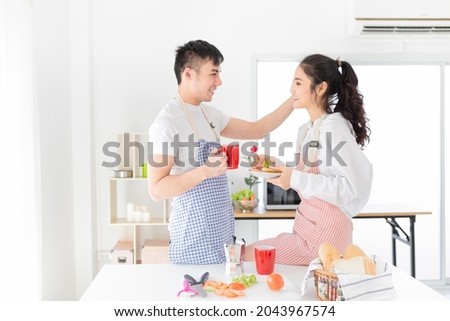asian man and asian woman eating meal in the morning, they have breakfast in kitchen room, he holding hot coffee cup, she holding sandwich with hand, they feeling happy and smile, happiness honeymoon 