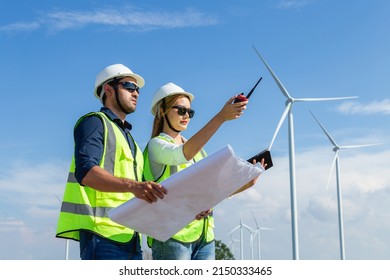 Asian Man And Woman Engineers Working On Site In Wind Turbine On The Background. Young People Engineers Working At Renewable Energy Farm