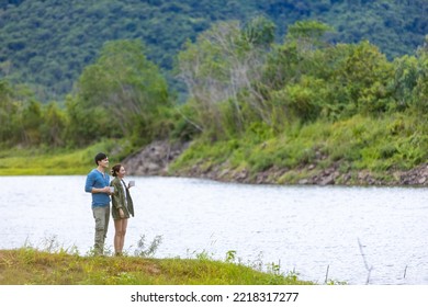 Asian Man And Woman Friends Drinking Coffee Together While Having Breakfast By The Lake In The Morning. Happy Family Enjoy Outdoor Lifestyle Camping In Forest Mountain On Summer Travel Vacation.