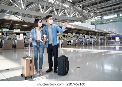 Asian Man And Woman Passengers Wear Face Mask Walk In Airport Terminal. Young Traveler Couple Stand In Boarding Gate During The COVID Pandemic. New Normal Lifestyle In Airport Transportation Concept.