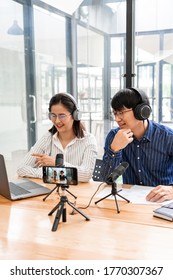 Asian Man And Woman Podcasters In Headphones Recording Content With Colleague Talking To Microphone And Camera In Broadcast Studio Together, Communication Technology And Entertainment Concept