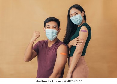 Asian Man And Woman Showing Shoulders After Getting A Vaccine. Happy Couple Showing Arm With Band-aids On After Vaccine Injection.