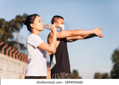 Asian Man And Woman Wearing Face Mask While Relaxing And Working Out On Street Together. She Drinking Water And Putting Off Mask And He Stretching Arms. Sport And Coronavirus Or Covid-19 Concept. 