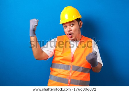 Asian man worker wearing safety helmet lookis happy celebrating his victory by clenching his fists against blue background