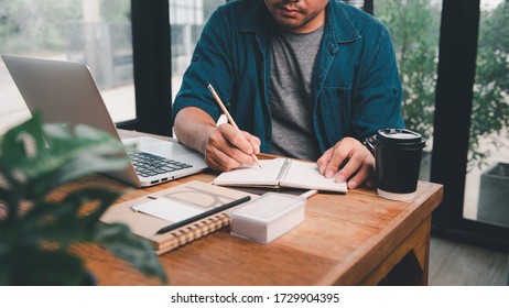 Asian Man Working At Desk With Notebook In Front And He Taking Notes On His Notebook.Home Working Ideas Table Setting Is Casual, With A Coffee Cup And A Tree To Purify The Air. And Equipment For Work.