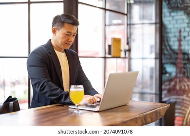 Asian Man Working On Laptop While Standing At The Bar With A Glass Of Beer. Concept Of Remote Work And Business At Pub With Alcohol. Man Wearing Formal Wear