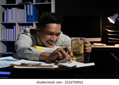 Asian Man Working Overtime Drinking Beer While Using Calculator In Office At Night.