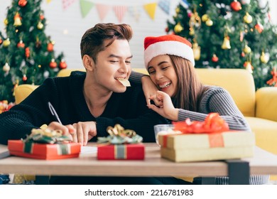 Asian man writing on a blank Christmas postcard with a pen while his girlfriend feeds him a potato chip. Couple sitting and writing Christmas card together at home during Christmas holiday. - Powered by Shutterstock