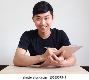 Asian Man Writing The Pen On Clipboard In Hand Look At Camera With Happy Smile Sit At The Desk,Register The Job Concept 