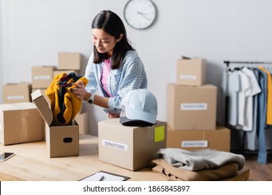 Asian Manager Of Charity Center Putting Clothes Near Boxes With Donations Lettering And Smartphone While Working In Charity Center 