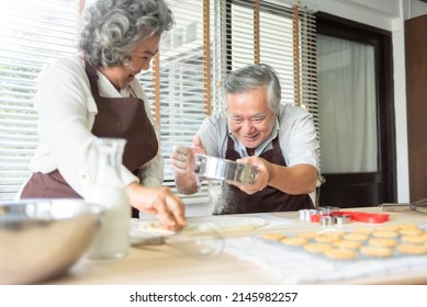 Asian Mature Adult Couple Cooking In Kitchen. Grandfather And Grandmother Sprinkling Dough With Flour Together.