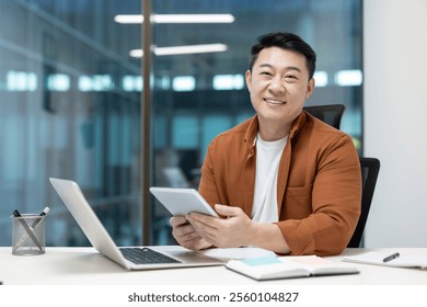 Asian mature businessman smiling while working on a tablet in a bright, professional office setting, representing modern technology usage and a productive work environment. - Powered by Shutterstock