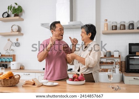 Asian mature couple playfully tease each other with grapes in kitchen, They talk and laugh with fun during eating fresh fruits with red grape, husband show thumb up