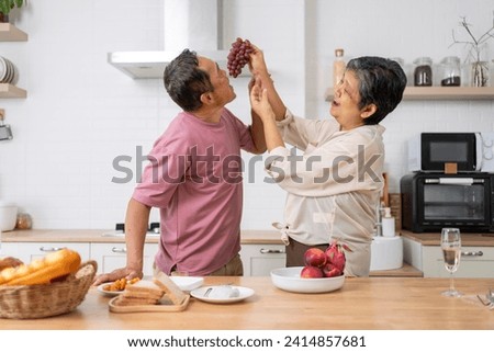 Asian mature couple playfully tease each other with grapes in kitchen, They talk and laugh with fun during eating fresh fruits with red grape