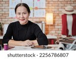 Asian mature female fashion designer is sitting at table, looking at camera and in studio, working with creative and sewing for dress design collection, professional boutique tailor SME entrepreneur. 