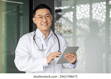 Asian mature male doctor in white coat using a tablet and smiling in a modern hospital or clinic setting, showcasing professional healthcare services. - Powered by Shutterstock