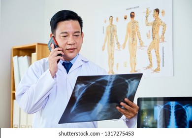 Asian Mature Male Neurologist In White Lab Coat Looking At X-ray Image And Talking On Mobile Phone At Office