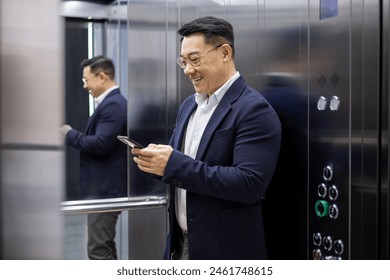 Asian mature man in a business suit smiling and using smartphone in an elevator. Professional enjoys the convenience of technology while commuting within an office building. - Powered by Shutterstock