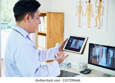 Asian Mature Neurologist Discussing X-ray Image With His Colleague Online On Digital Tablet At Hospital