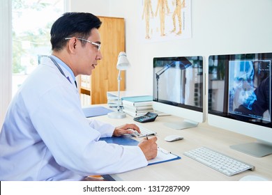 Asian Mature Neurologist In Eyeglasses Examining X-ray Image On Computer Monitor And Making Notes On Clipboard While Working At His Workplace