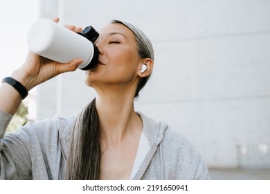 Asian Mature Woman In Earphones Drinking Water During Workout Outdoors