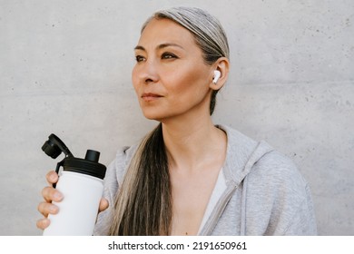 Asian Mature Woman In Earphones Drinking Water During Workout Outdoors