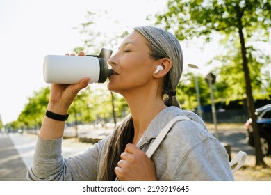 Asian Mature Woman In Earphones Drinking Water During Workout Outdoors