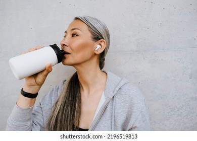 Asian Mature Woman In Earphones Drinking Water During Workout Outdoors