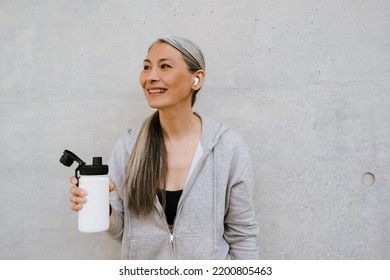 Asian Mature Woman In Earphones Drinking Water During Workout Outdoors